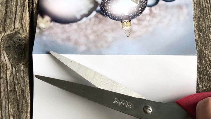 Eine Hand schneidet mit einer Schere eine Weihnachtskarte entlang der Faltlinie auf einem Holztisch.