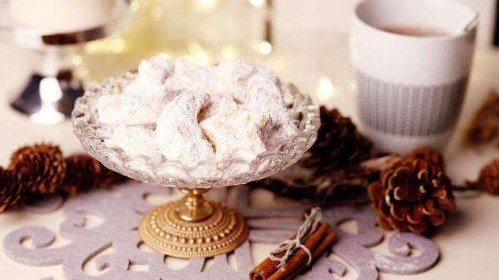 Eine Glasschale mit Weihnachtspl&auml;tzchen steht auf einem dekorativen Tisch, umgeben von Tannenzapfen und einer Tasse.