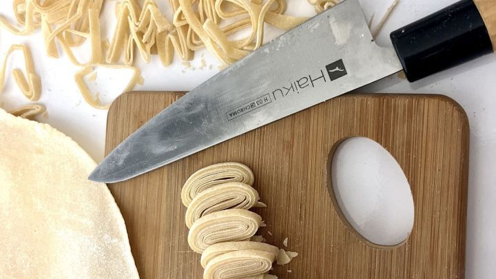 Frische Nudeln liegen auf einem Brett, w&auml;hrend ein Messer sie in gleichm&auml;&szlig;ige Streifen schneidet, um leckere Speisen zuzubereiten.