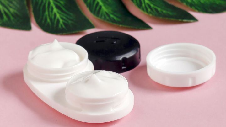 Ein Kontaktlinsenbeh&auml;lter mit zwei F&auml;chern zeigt die darin befindliche Creme, umgeben von gr&uuml;nen Bl&auml;ttern auf rosa Hintergrund.