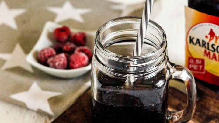 Ein Glas mit dunklem Getr&auml;nk und einem Strohhalm steht auf einem Holzteller, umgeben von Himbeeren und einer Flasche.