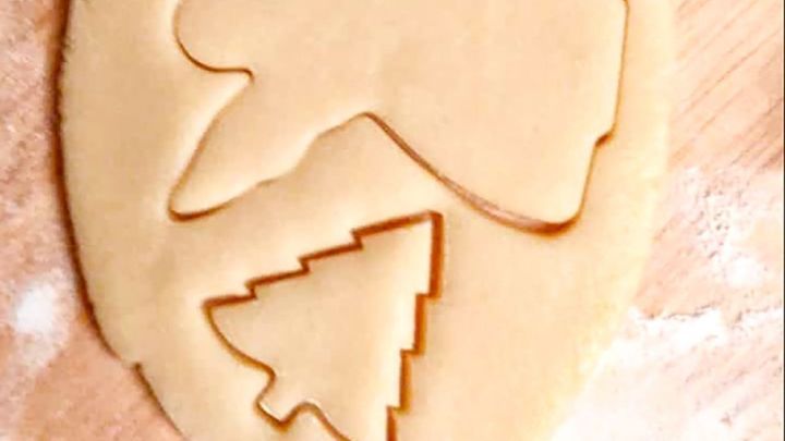 Ein Ausstecher formt einen Engel und einen Weihnachtsbaum aus einem ausgerollten Pl&auml;tzchenteig auf einem Holzbrett.