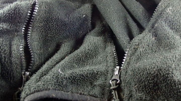 Eine schwarze, flauschige Jacke mit zwei offenen Rei&szlig;verschl&uuml;ssen liegt zusammengefaltet auf einem Hintergrund.