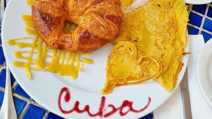 Ein Croissant und eine herzhafte Omelette auf einem Teller mit der Aufschrift &bdquo;Cuba&ldquo; vor einem blauen Mosaiktisch.