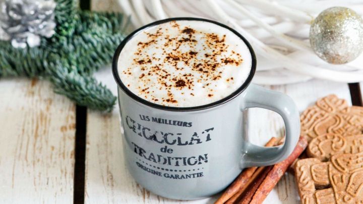 Ein grauer Becher mit Cappuccino steht auf einem rustikalen Tisch, umgeben von Weihnachtsdekoration und einer Laterne.