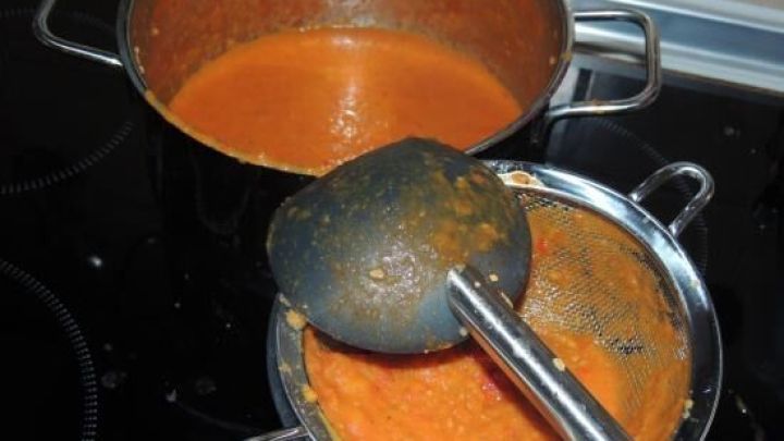 Eine silberne Kasserolle mit einer leuchtend roten Knoblauch-Tomaten-Paprika-Suppe wird mit einem Sch&ouml;pfl&ouml;ffel angerichtet.