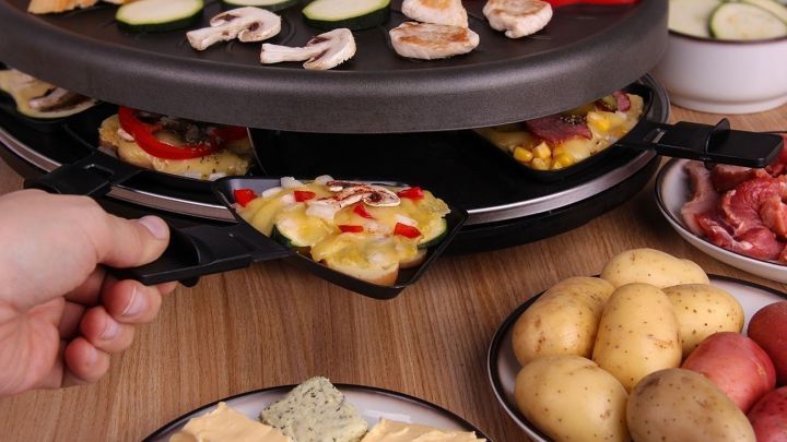 Ein Hand greift nach einem Pf&auml;nnchen vom Raclettegrill, w&auml;hrend Gem&uuml;se und Fleisch auf dem Grill gargezogen werden.