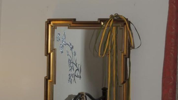 Ein goldener Rahmen um einen Spiegel h&auml;ngt an der Wand, geschm&uuml;ckt mit einer gelben Schleife, vor einem hellen Hintergrund.