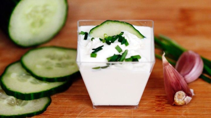 Tzatziki in einem Glas mit Gurkenscheiben und feingehacktem Schnittlauch, auf einem Holzbrett angerichtet.