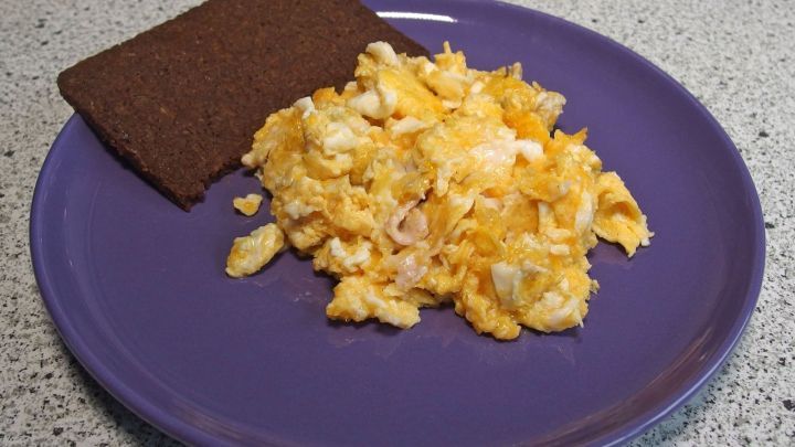Ein lila Teller zeigt R&uuml;hrei, das neben einer Scheibe dunklem Brot angerichtet ist, auf einer grauen Tischoberfl&auml;che.
