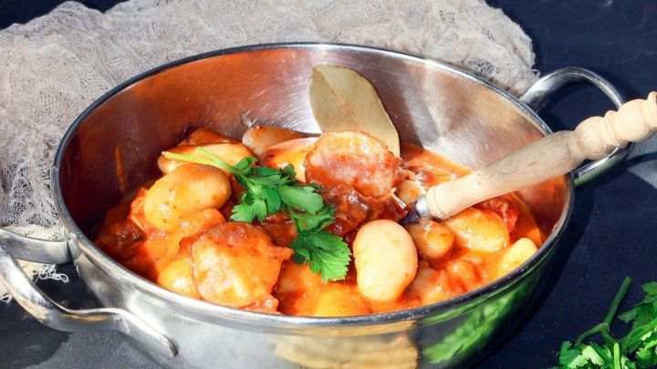 Ein gl&auml;nzender Topf mit wei&szlig;em Bohneneintopf steht auf einem dunklen Untergrund. Eine Holzl&ouml;ffel r&uuml;hrt die Mischung.