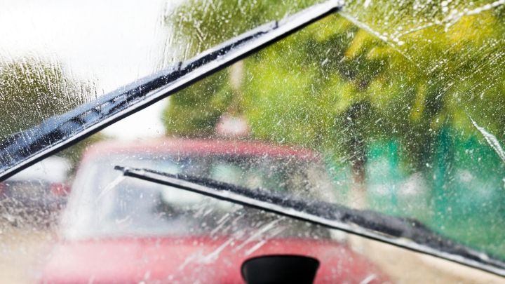 Scheibenwischer reinigen die nasse Windschutzscheibe eines Autos, w&auml;hrend im Hintergrund ein rotes Fahrzeug zu sehen ist.