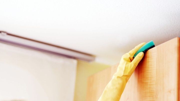 Eine Hand in gelben Gummi-Handschuhen reinigt den oberen Rand eines K&uuml;chenschranks, um fettigen Dreck zu entfernen.