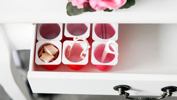 Im offenen Schublade sind leere Joghurtbecher angeordnet, w&auml;hrend rosa Blumen die Oberfl&auml;che dekorieren.