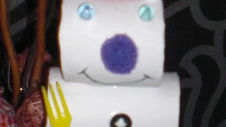 Ein Schneemann aus Klopapierrollen l&auml;chelt mit buntem Hut und gelbem Besteck in einem festlich dekorierten Raum.