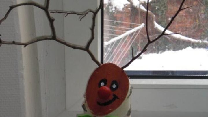 Ein Rentier aus einem Baumstamm steht mit Astgeweihen und einem l&auml;chelnden Gesicht auf einem Fensterbrett in einer verschneiten Umgebung.