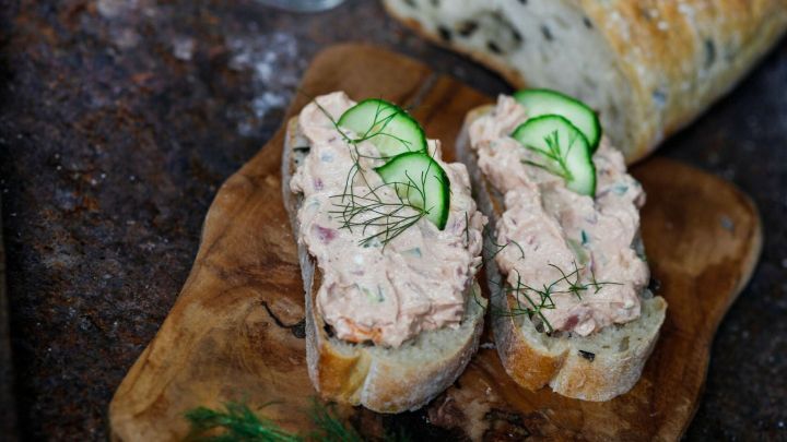 Auf einer Holzplatte liegt frisches Brot, belegt mit Frischk&auml;se und Gurkenscheiben, umgeben von Blumen und einem Brotlaib.