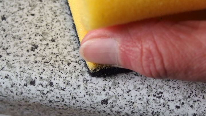 Eine Hand h&auml;lt einen Schwamm, der Sekundenkleber von einer grauen Arbeitsplatte entfernt, w&auml;hrend der Raum hell beleuchtet ist.