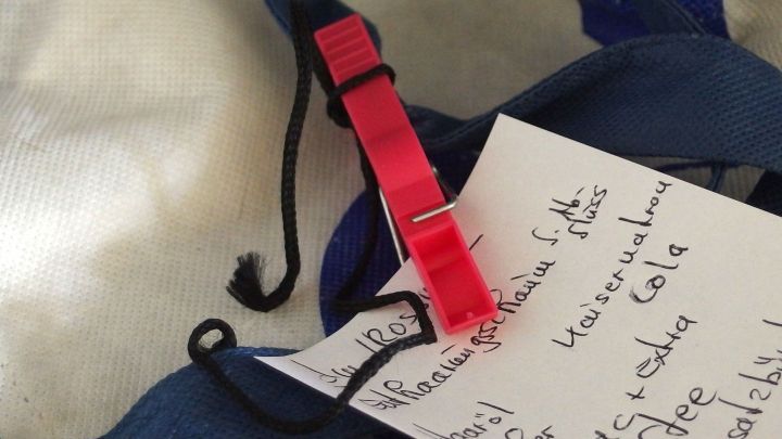 Eine rote Klammer h&auml;lt einen Einkaufszettel mit Waren zum Einkaufen auf einem blauen Stoff in einer neutralen Umgebung.