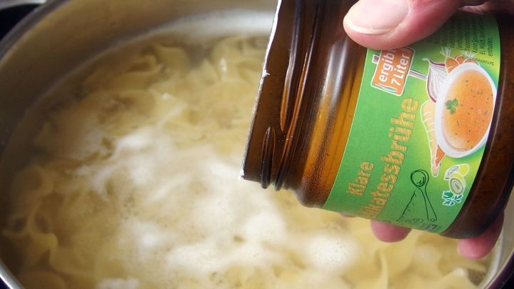 Eine Hand h&auml;lt ein Glas mit Gew&uuml;rzen &uuml;ber einen Topf mit sprudelnden Nudeln, die auf dem Herd gekocht werden.