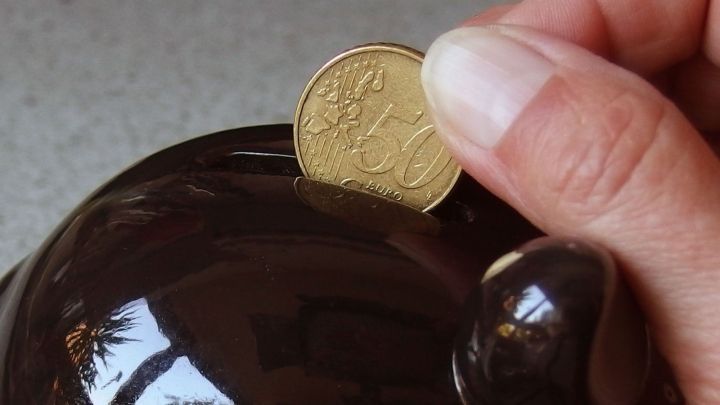 Eine Hand steckt eine M&uuml;nze in ein braunes Sparschwein, um Kindern den Umgang mit Geld n&auml;her zu bringen.