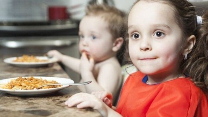 Ein kleines M&auml;dchen in einem roten Outfit sitzt vor einem Teller Essen, w&auml;hrend ihr Bruder interessiert zuschaut.