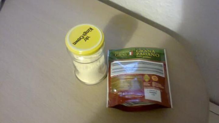 Ein Schraubglas mit Parmesan steht auf einem Tisch neben einer Verpackung, bereit zum Umf&uuml;llen f&uuml;r die Aufbewahrung.