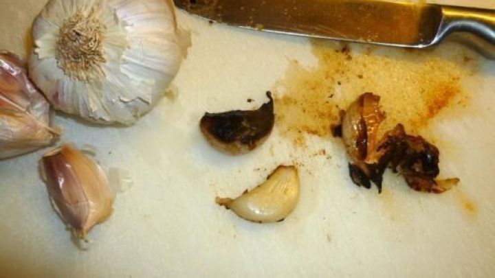 Knoblauchzehen liegen auf einem Schneidebrett, w&auml;hrend eine Klinge daneben geviertelte, leicht angebratene Zehen zeigt.