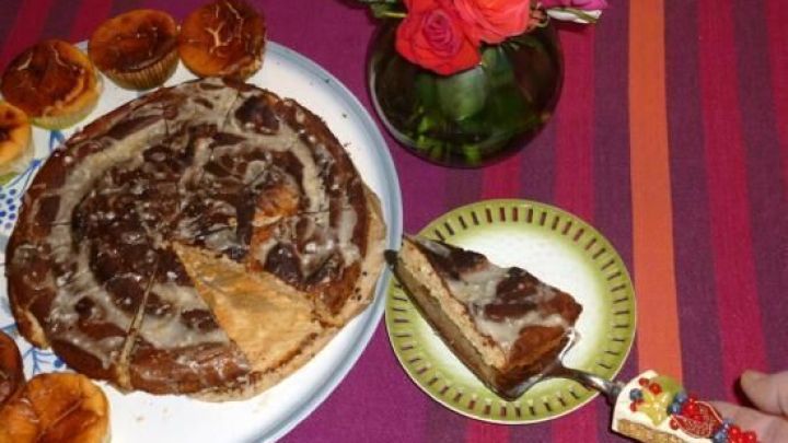 Ein Zitrus-Mandel-Kuchen liegt auf einem Teller, w&auml;hrend ein St&uuml;ck auf einen kleinen Teller serviert wird, umgeben von Geb&auml;cken und einer Blumenvase.