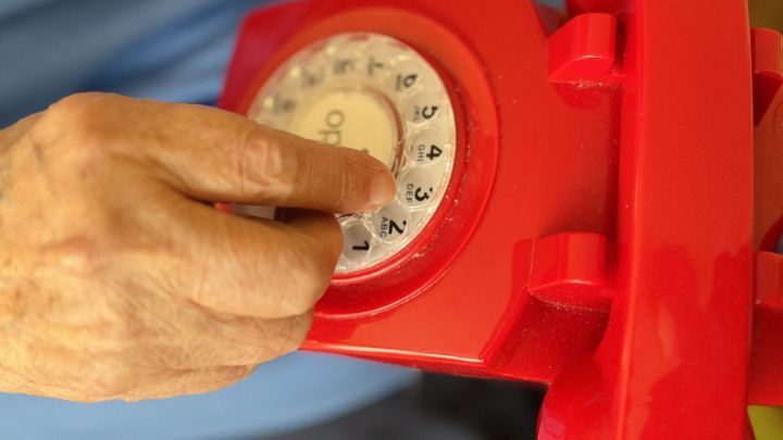 Eine Hand dreht eine Nummer auf einem roten W&auml;hlscheibentelefon, w&auml;hrend jemand in einem hellen Raum sitzt.