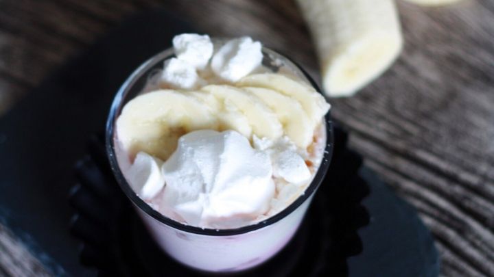 Ein Dessertbecher mit Sahne und Bananenscheiben, umgeben von frischen Bananen auf einem Holztisch.