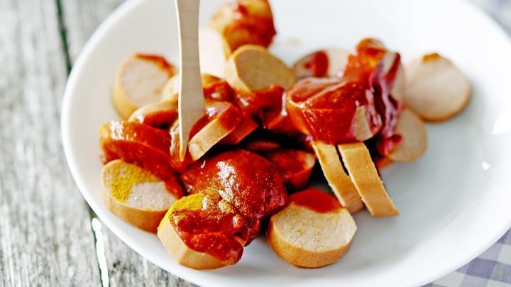 Auf einem wei&szlig;en Teller sind geschnittene Wurstst&uuml;cke mit roter Sauce angerichtet, umgeben von einem karierten Tuch.