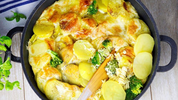 Ein goldbraun gebackener Nudelauflauf mit Kartoffeln und Brokkoli in einer schwarzen Pfanne steht auf einem Holzuntergrund.