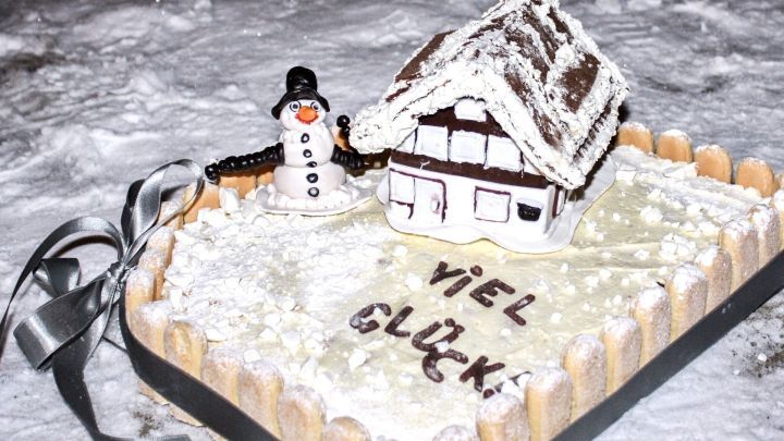Ein sch&ouml;n dekorierter Kuchen mit einem kleinen H&auml;uschen und einem Schneemann, umgeben von Schnee, steht festlich auf einem Tisch. Der Text &bdquo;VIEL GL&Uuml;CK&ldquo; ist darauf geschrieben.