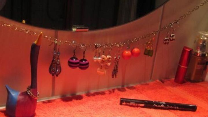 Ohrringe h&auml;ngen an einer Kette &uuml;ber einem Badspiegel, w&auml;hrend ein roter Schminkuntergrund und Kosmetikartikel sichtbar sind.