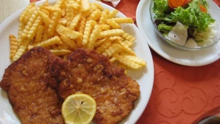 Schnitzel mit Zitronenscheibe und Pommes frites auf einem Teller, daneben ein frischer Salat, auf orangem Tischtuch serviert.