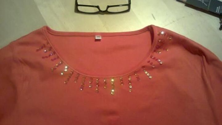 Ein rotes Shirt mit funkelnden Pailletten sitzt auf einem Tisch, umgeben von Brille und N&auml;hzubeh&ouml;r.