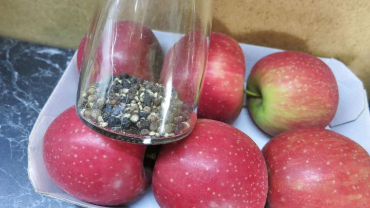 Ein Glas Pfeffer wird &uuml;ber eine Schale mit gl&auml;nzenden roten &Auml;pfeln gehalten, die auf einer dunklen Oberfl&auml;che liegen.