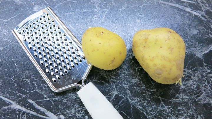 Eine Reibe liegt neben zwei rohen Kartoffeln auf einer dunklen, marmorierten Oberfl&auml;che, bereit zum Raspeln.