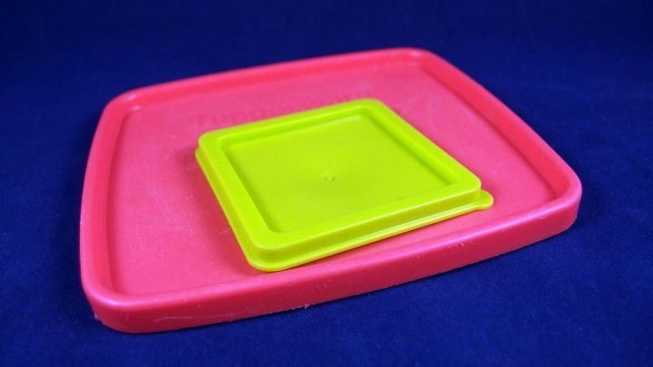 Ein rosa Tupperdeckel liegt offen auf einem blauen Hintergrund, w&auml;hrend ein kleiner, gr&uuml;ner Silikondeckel darauf platziert ist.