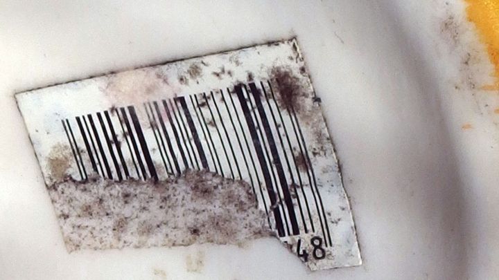Ein schmutziges Klebeetikett mit einem Barcode liegt auf einer wei&szlig;en Oberfl&auml;che und zeigt Spuren von Ablagerungen.