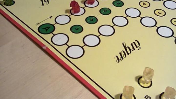 Auf einem Brettspiel f&uuml;r Kinder bewegen sich gr&uuml;nes und rotes Spielzeug auf einem bunten Spielfeld in einem gem&uuml;tlichen Raum.