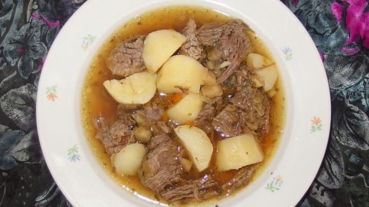 Ein Rindfleischtopf mit Thymian und Kartoffeln liegt in einer wei&szlig;en Sch&uuml;ssel auf einem bunten Tisch.