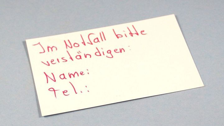 Ein kleiner Zettel liegt auf einer grauen Oberfl&auml;che, auf dem steht: &bdquo;Im Notfall bitte verst&auml;ndigen. Name: ___ Tel.: ___&ldquo;.