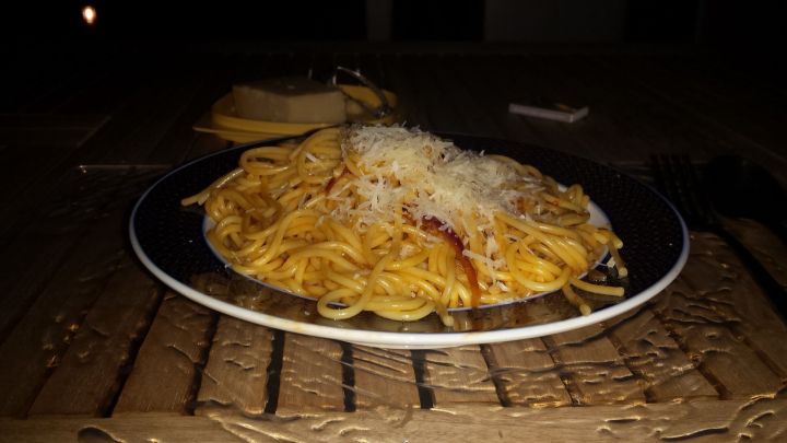 Ein Teller Spaghetti mit Tomatenso&szlig;e und geriebenem K&auml;se steht auf einem Holztisch im Freien, daneben liegt ein St&uuml;ck K&auml;se.