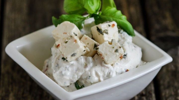 Eine cremige Schafsk&auml;secreme mit St&uuml;ckchen, garniert mit frischem Basilikum, steht in einer Sch&uuml;ssel auf einem Holzuntergrund.