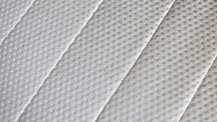 Eine alte Matratze mit abgenutztem, strukturiertem Bezug liegt auf einem Bett, umgeben von neutralem Schlafzimmerlicht.