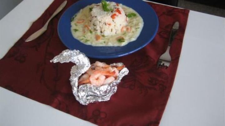 Ein festlicher Alufolienbeh&auml;lter in Schwansform pr&auml;sentiert Garnelen neben einem Teller mit Reis und Sauce auf einem Tisch.