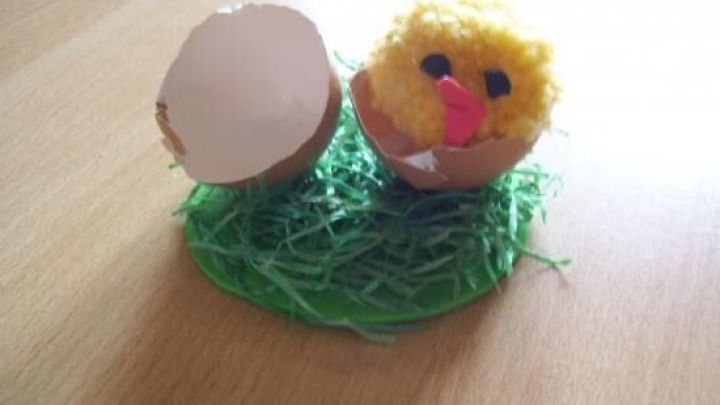 Ein gelbes, flauschiges K&uuml;ken sitzt auf gr&uuml;nem Gras und schaut aus einer zerbrochenen Eierschale, fr&ouml;hliche Osterdeko.