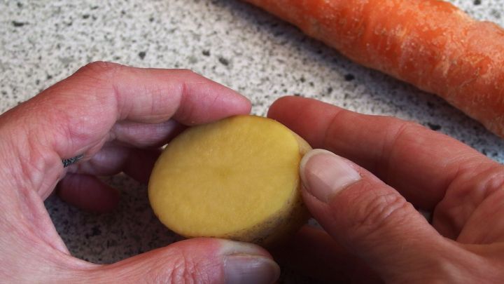 Eine Hand h&auml;lt eine halbgesch&auml;lte M&ouml;hre, w&auml;hrend die andere Hand eine Kartoffel greift, alles auf einer K&uuml;chenoberfl&auml;che.